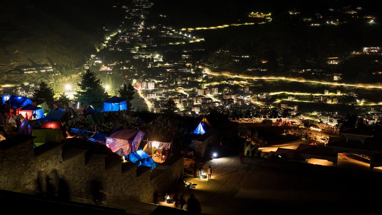 Nighttime view of a Himalayan hillside town glowing with winding street lights, colorful tented shelters in the foreground, and scattered buildings illuminating the valley below.
