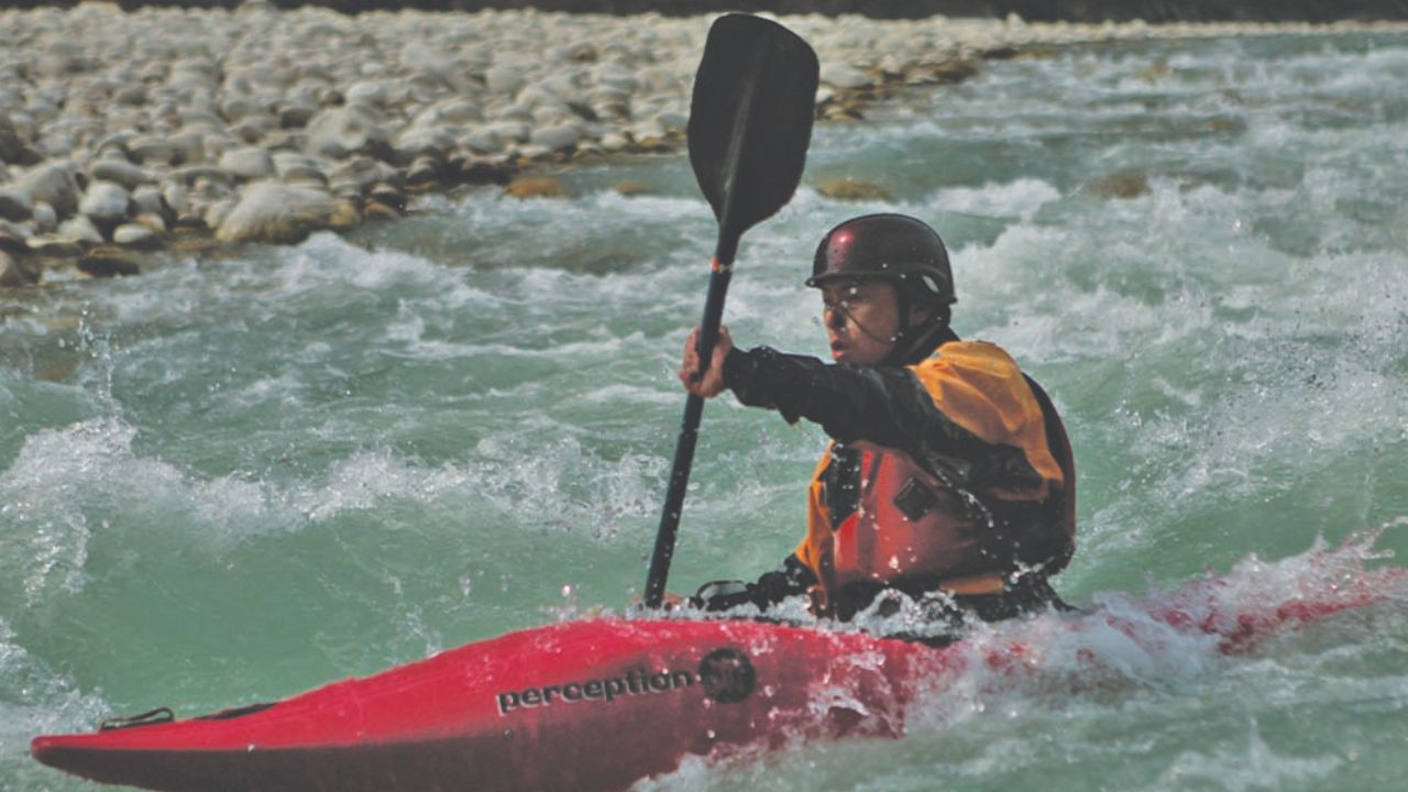 Kayaker navigating whitewater rapids on a fast-flowing mountain river while wearing a helmet and safety gear.
