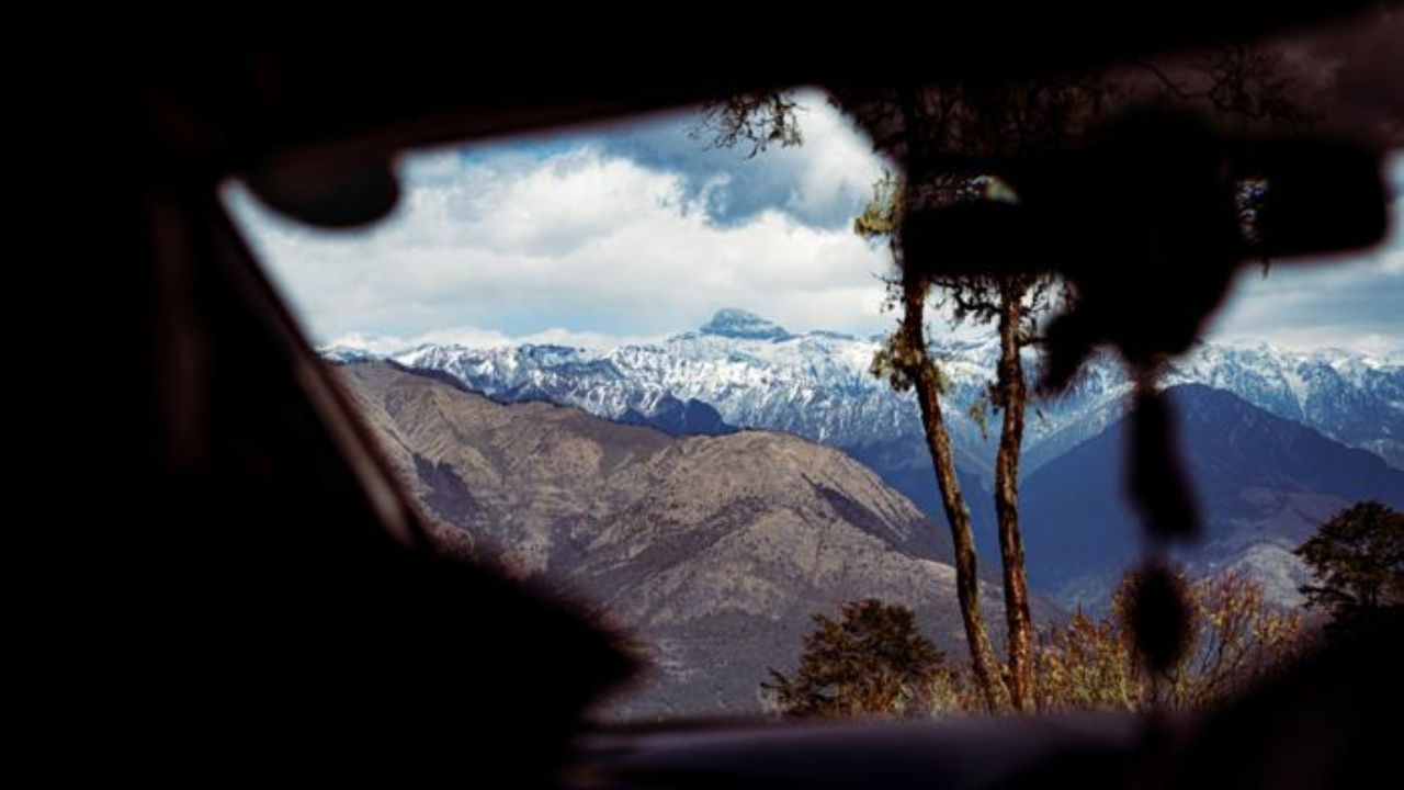Snowy mountains seen through a car windshield, framed by the dark interior and a tree.